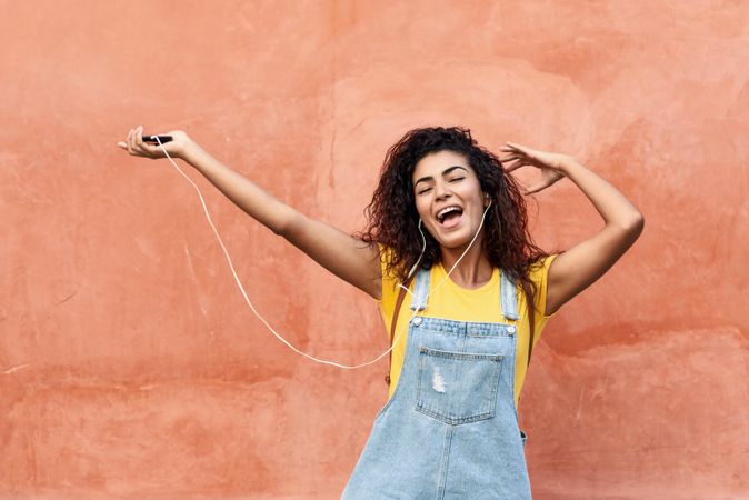 Dancing and singing Arab woman with curly hairstyle using phone and headphones with arms in the air