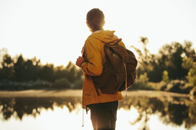 Young man wearing a backpack standing near a lake and looking at the evening sun