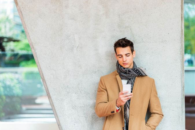 Man in fall coat texting while leaning back on cement pillar