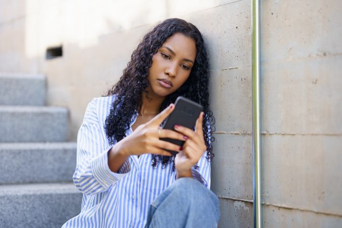 A young woman is focused on her smartphone while sitting on steps, enjoying her leisure