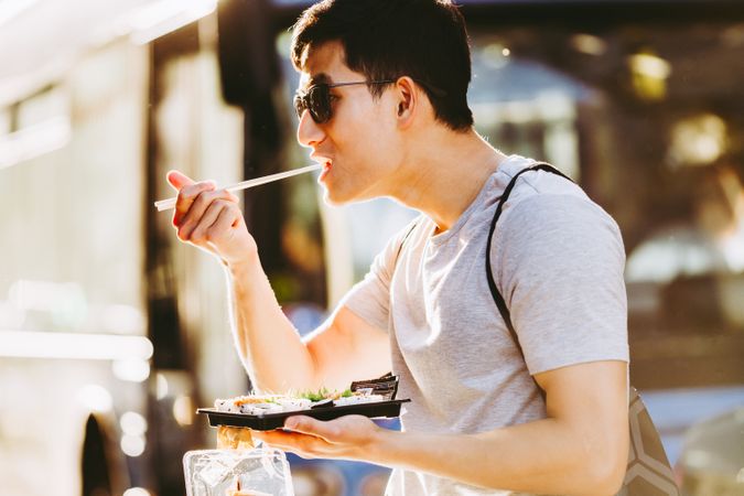Asian man eating takeaway food with chopsticks on city street