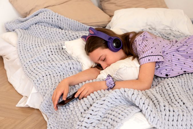 Teenage girl asleep in bed with headphones and smartphone.