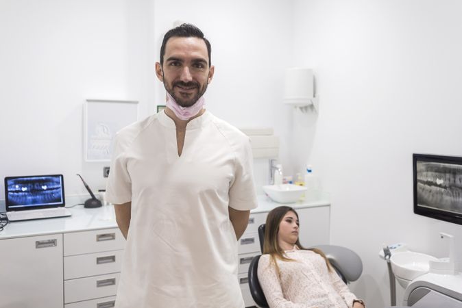 Smiling male dentist pictured in his office with teenage patient in background