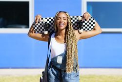 Happy female in denim overalls standing with skateboard behind head in front of blue building