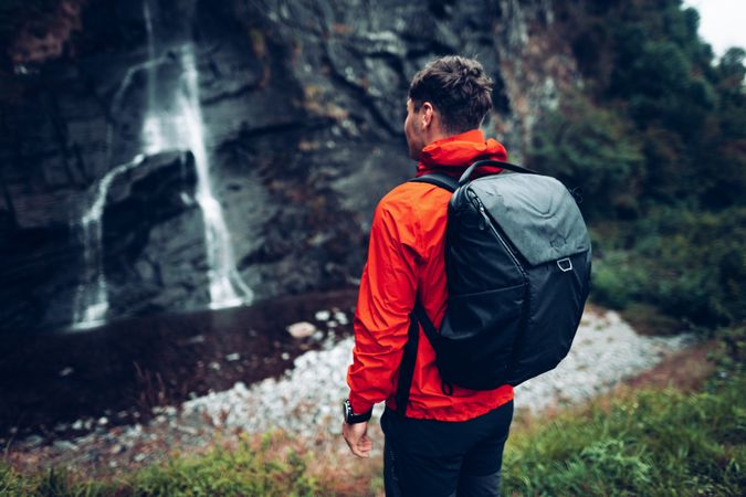 Back view of a man in red jacket with backpack standing facing waterfall during daytime