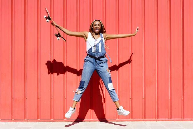 Woman smiling and jumping in front of a red wall with skateboard