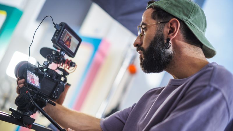 Focused Male Cinematographer With Beard Operates Professional Video Camera On Film Set, Capturing Dynamic Footage For A Documentary Or Tv Show, Surrounded By Colorful Set Design.