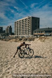 Boy pushing bike on sand in front of building bxZwa5