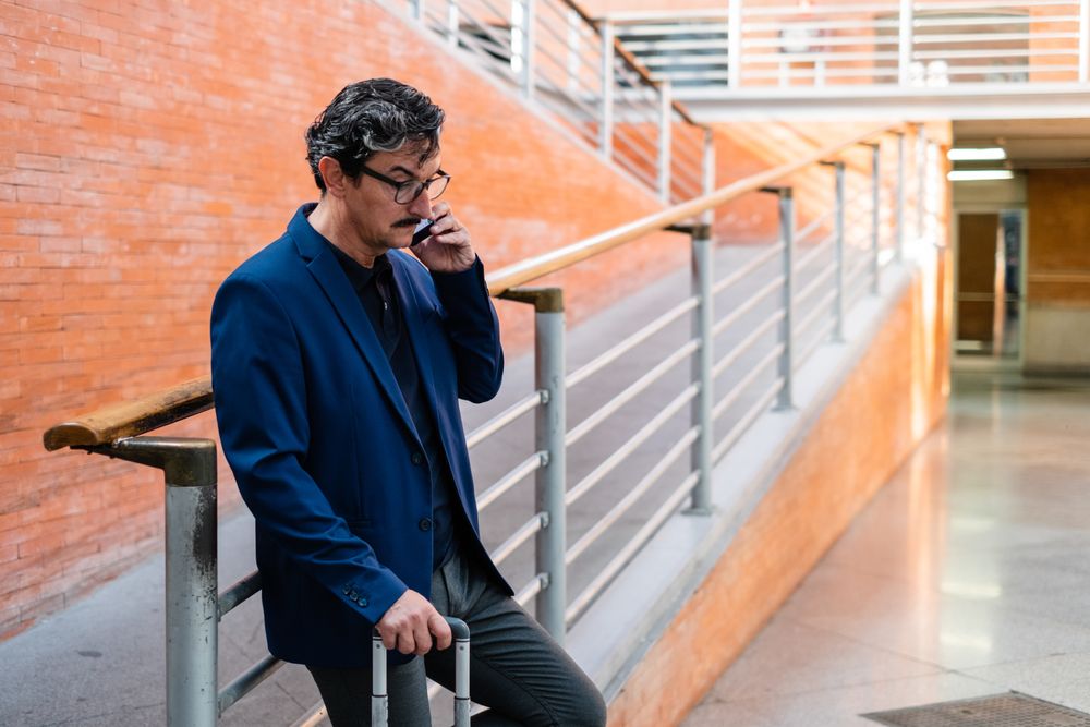 Puzzled mature man standing with baggage and talking on smartphone