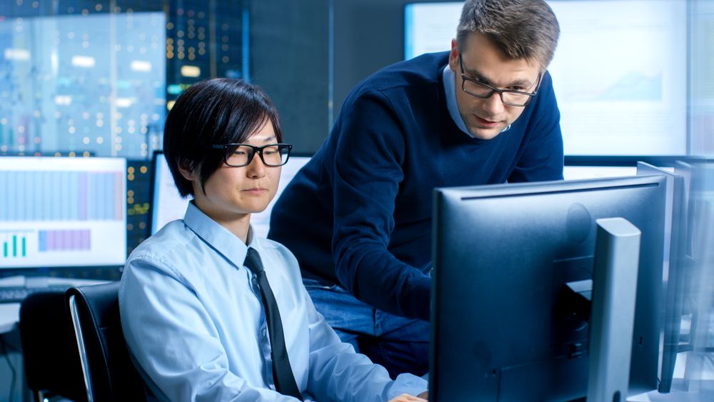 In the Data Mining Center Chief Statistician Consults His Young Assistant sits at His Workstation Surrounded with Monitors Displaying Data and Graphs.