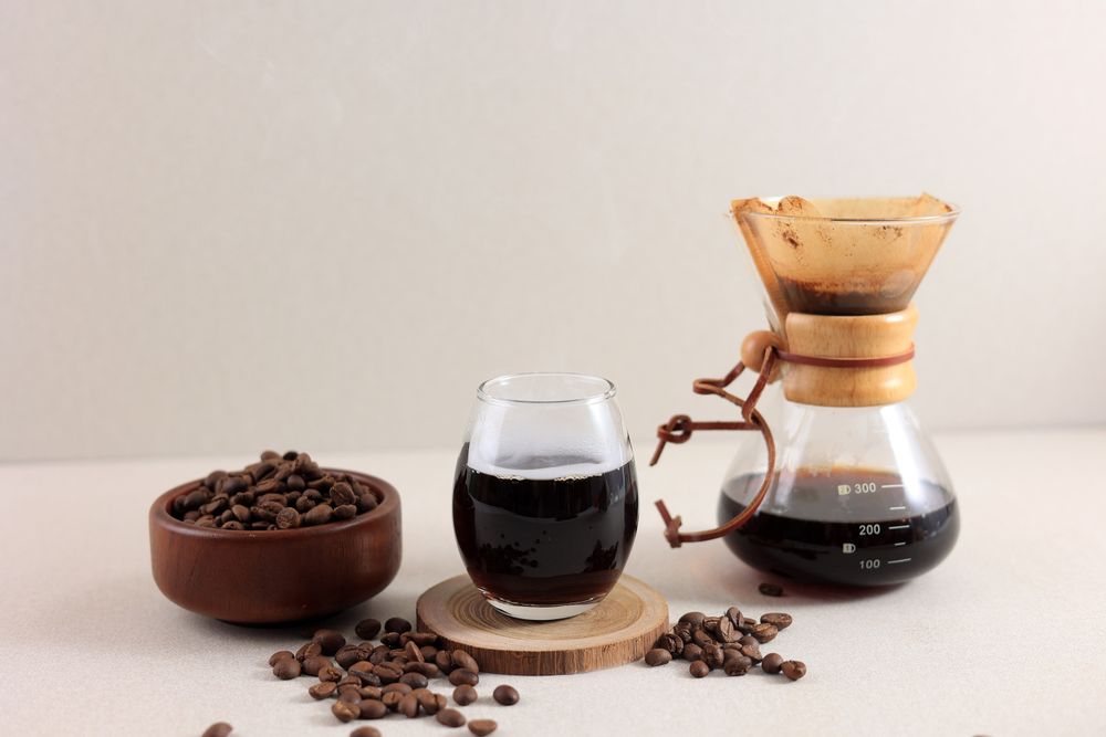 Glass Chemex Coffeemaker with Tasty Drip Coffee and Beans on Cream Table