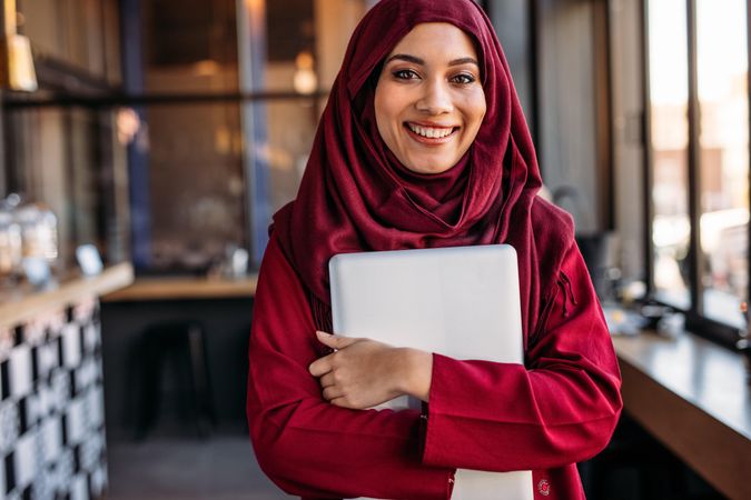 Portrait of happy young woman wearing hijab holding laptop computer looking at camera and smiling