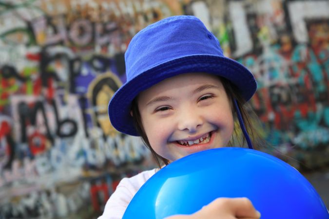 Close up portrait of little girl in blue hat holding a blue playball standing in front of graffiti