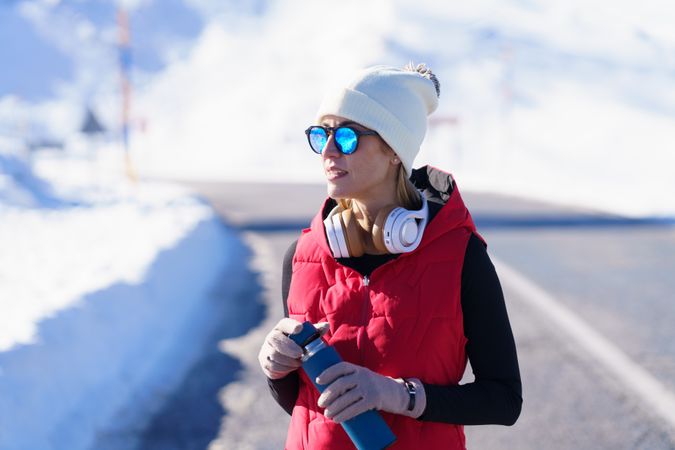 Female in warm clothes and sunglasses holding thermos bottle in snowy mountains