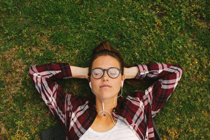 Top view of woman wearing eyeglasses lying on grass with hands behind head and eyes closed