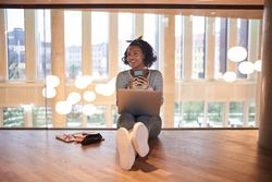 Woman sitting on floor smiling and working on her laptop with her cell phone