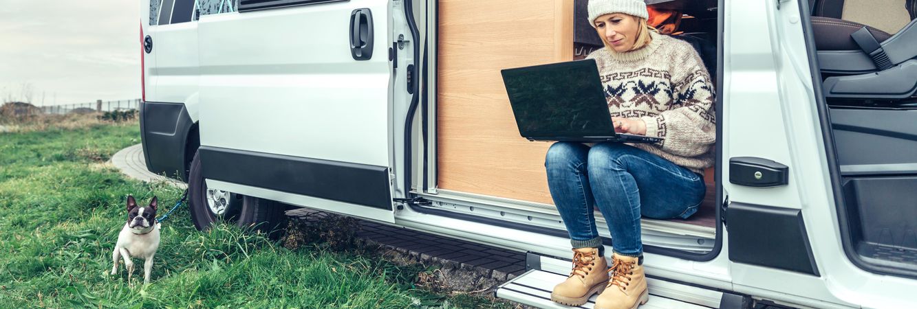Female in sweater working remotely sitting on step of van while on a road trip, wide