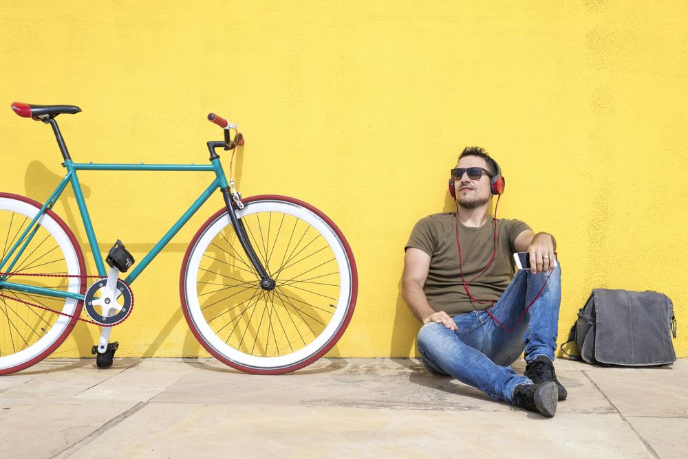 Male looking up in front of yellow wall next to bike with bike and listening to music