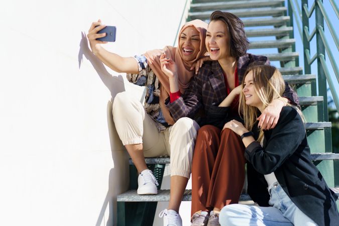 Multi-ethnic women friends in stylish outfits taking selfie on phone on metal stairs outside