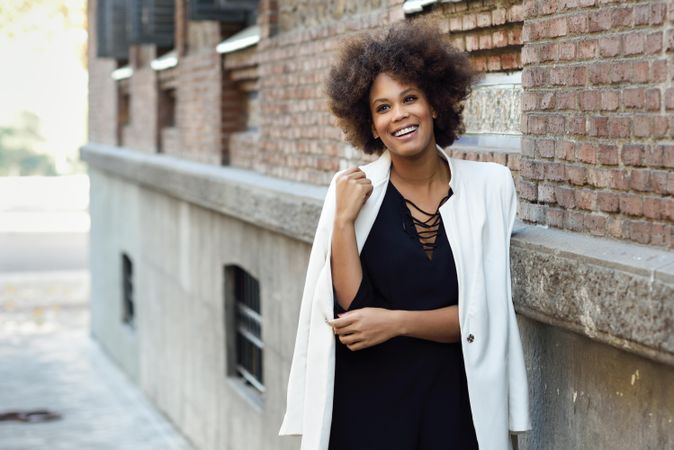 Smiling female in dress with blazer draped over shoulders standing next to outdoor wall