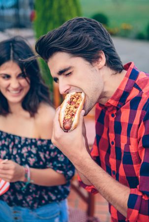 Young man eating hot dog and woman laughing in background