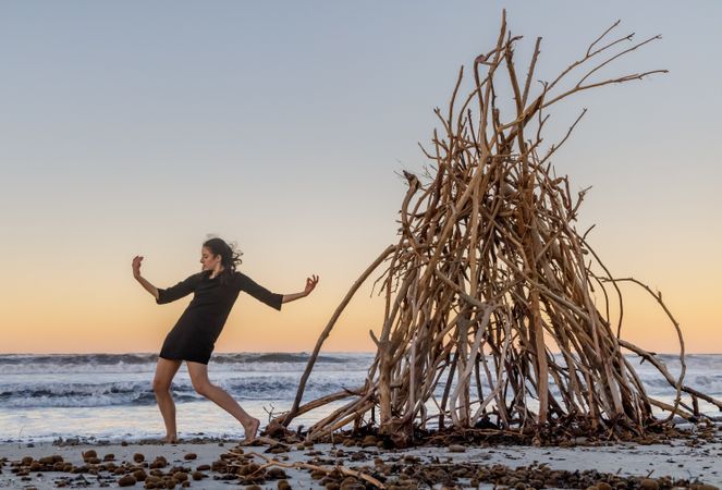Young woman dancing on shore near water and wooden twigs placed for campfire