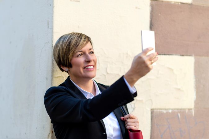 Smiling woman wearing a blazer and taking photo with phone outside