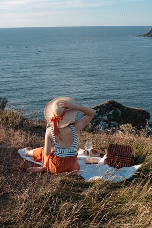 Woman with glass of drink on picnic mat looking at sea and mountains