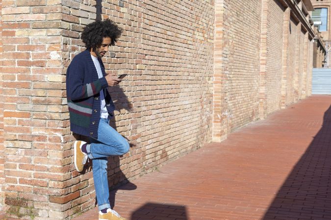 Black male using his smartphone while leaning on a bricked wall outdoors on sunny day