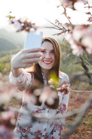 Young happy woman taking selfie on smartphone near trees on hill