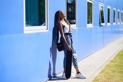 Female in denim overalls standing with skateboard while leaning on blue wall