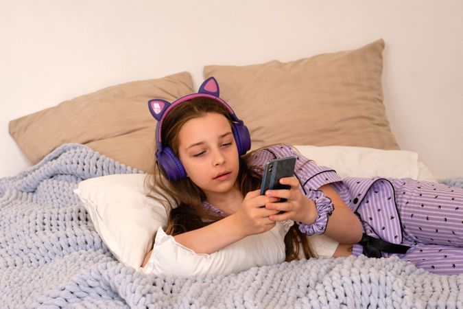 Girl lying in bed with headphones, using smartphone.