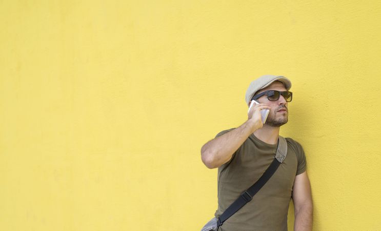Male in hat and sunglasses standing next to yellow wall and speaking on phone, copy space