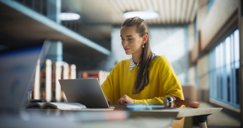 Female Student Working on Her College Degree Thesis on a Laptop Computer in Library. Young Stylish Female in a Yellow Jumper Studying an Online Course Alone in a Reading Hall