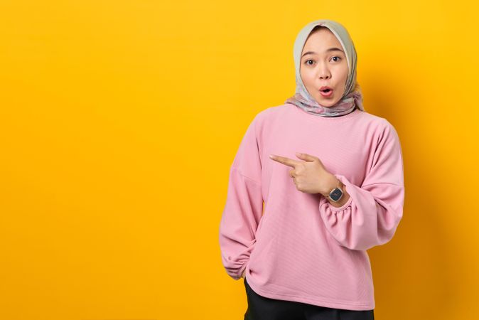 Surprised Muslim woman in scarf and pink sweater pointing in studio shoot