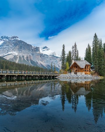 Brown wooden house near lake under snow covered mountain with reflection on water