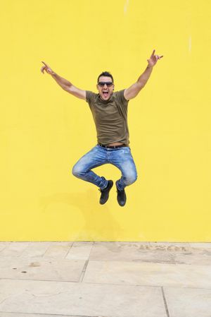Excited male standing outside jumping in front of yellow wall with arms up