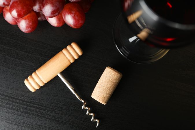 Grapes, corkscrew, cork and wine glass on dark table, top view