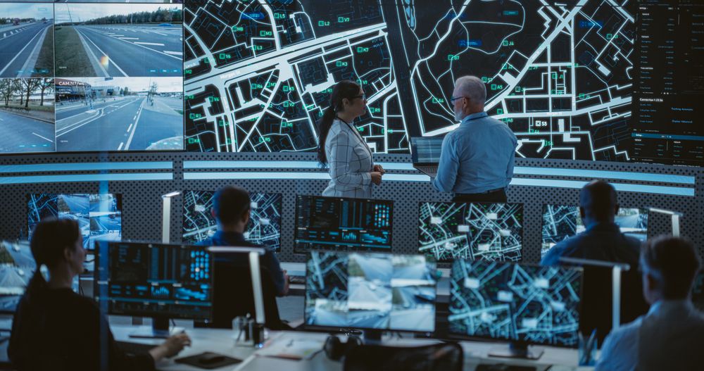 Two Multiethnic Software Engineers Talking Next to a Big Screen in a Modern Monitoring Office with Live CCTV Footage with Traffic Situation.