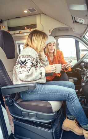 Female friends sitting in parked van having coffee and checking social media on phone, vertical
