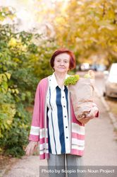 Older woman in pink cardigan holding grocery bag standing beside trees 4dWVL5