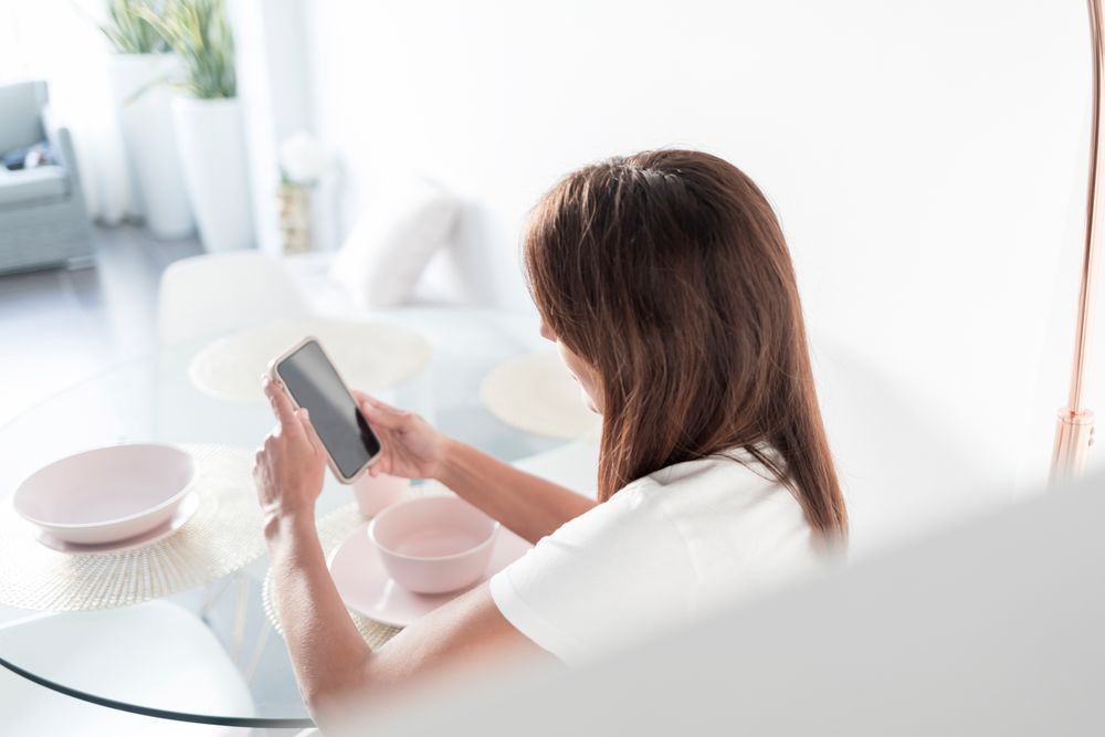 Back of female sitting at sunny breakfast table texting on phone in front of cereal bowl