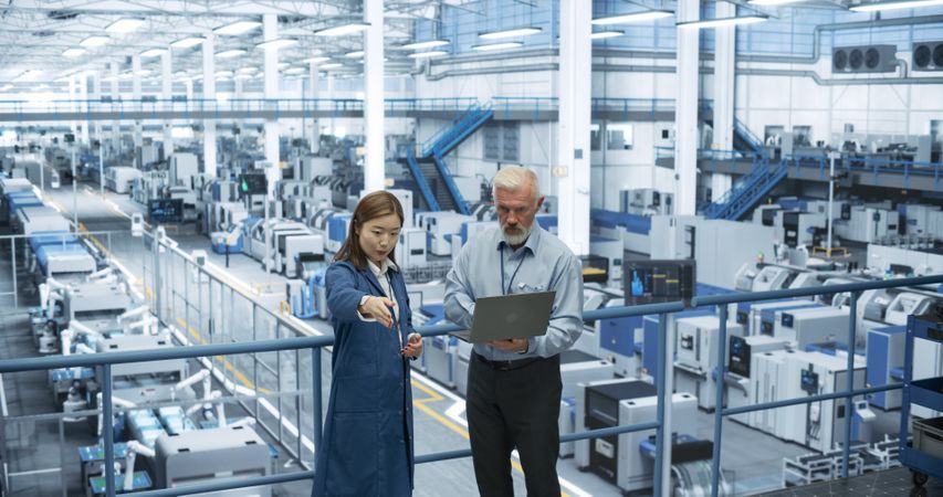 Mature Engineer and Young Japanese Technician Having a Conversation and Using Laptop Computer. Specialists Monitoring, Analyzing Conditions at a Modern Electronics Factory with Automated Robots