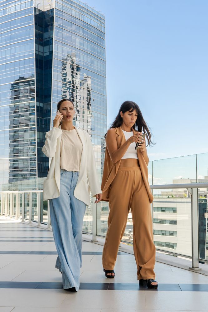 Two businesswomen are walking on a glass walkway. One of them is talking on the phone, and the other is sending a message.
