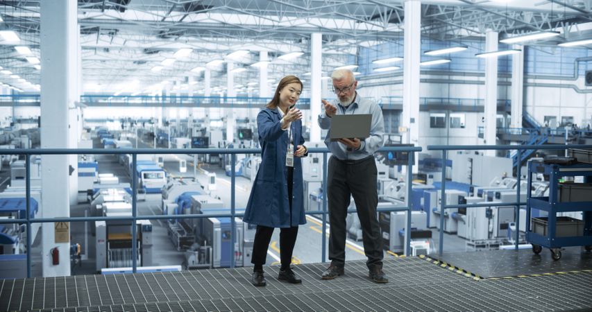 Two Diverse Engineers Standing on a Platform, Using Laptop Computer at an Electronics Factory. Machines are Producing Electronic Chips, Specialists Monitoring the Progress on Online Software