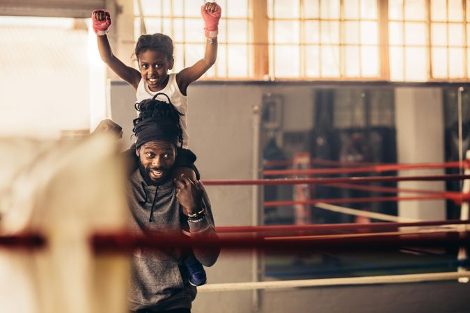 Girl in boxing gym sitting on the shoulders of coach with raised arms celebrating victory
