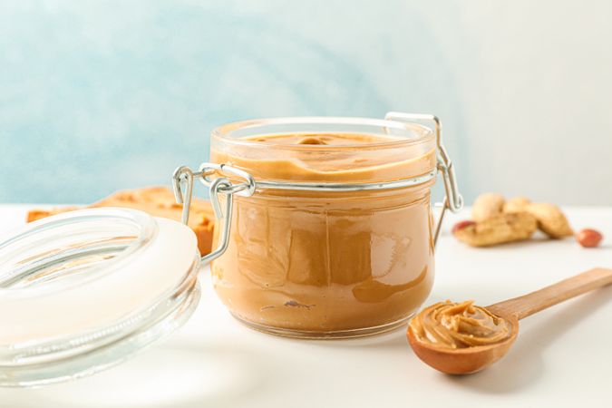 Composition with peanut butter sandwich, glass jar, peanut and spoon on light table against light background, space for text and closeup