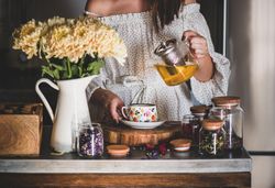Woman in pouring tea in floral cup and saucer from glass teapot, close up