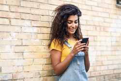 Smiling Arab woman texting with her smart phone leaning on brick wall