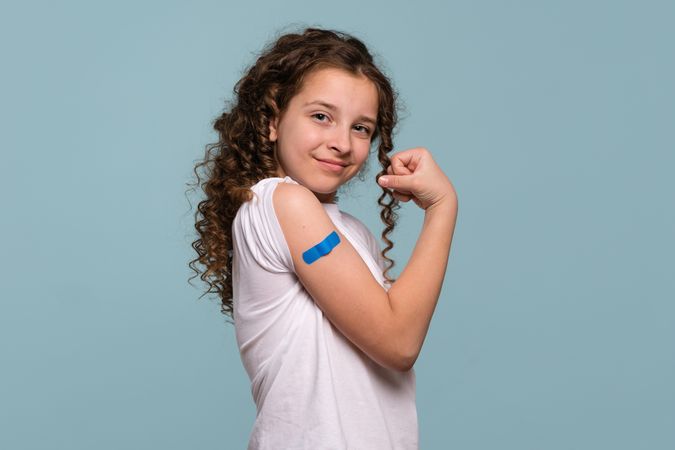 Girl flexing arm with blue bandage after receiving vaccine shot.